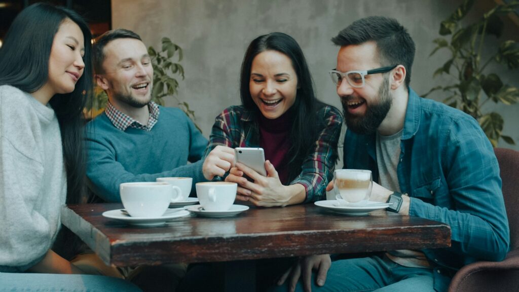 Four friends laughing while watching a video on a smartphone in a cafe